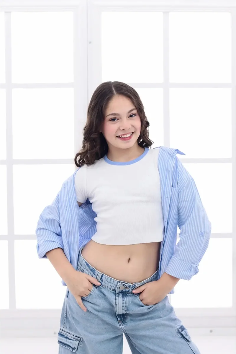 Niña con camisa celeste sonriendo frente a una ventana blanca