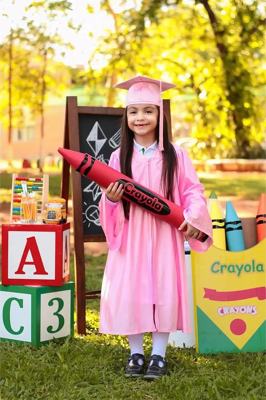 Niña con toga rosada y un crayón gigante en un set escolar al aire libre